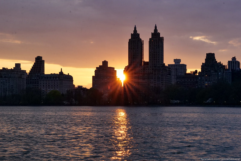 A view of the setting sun between buildings, as seen across the Central Park reservoir.