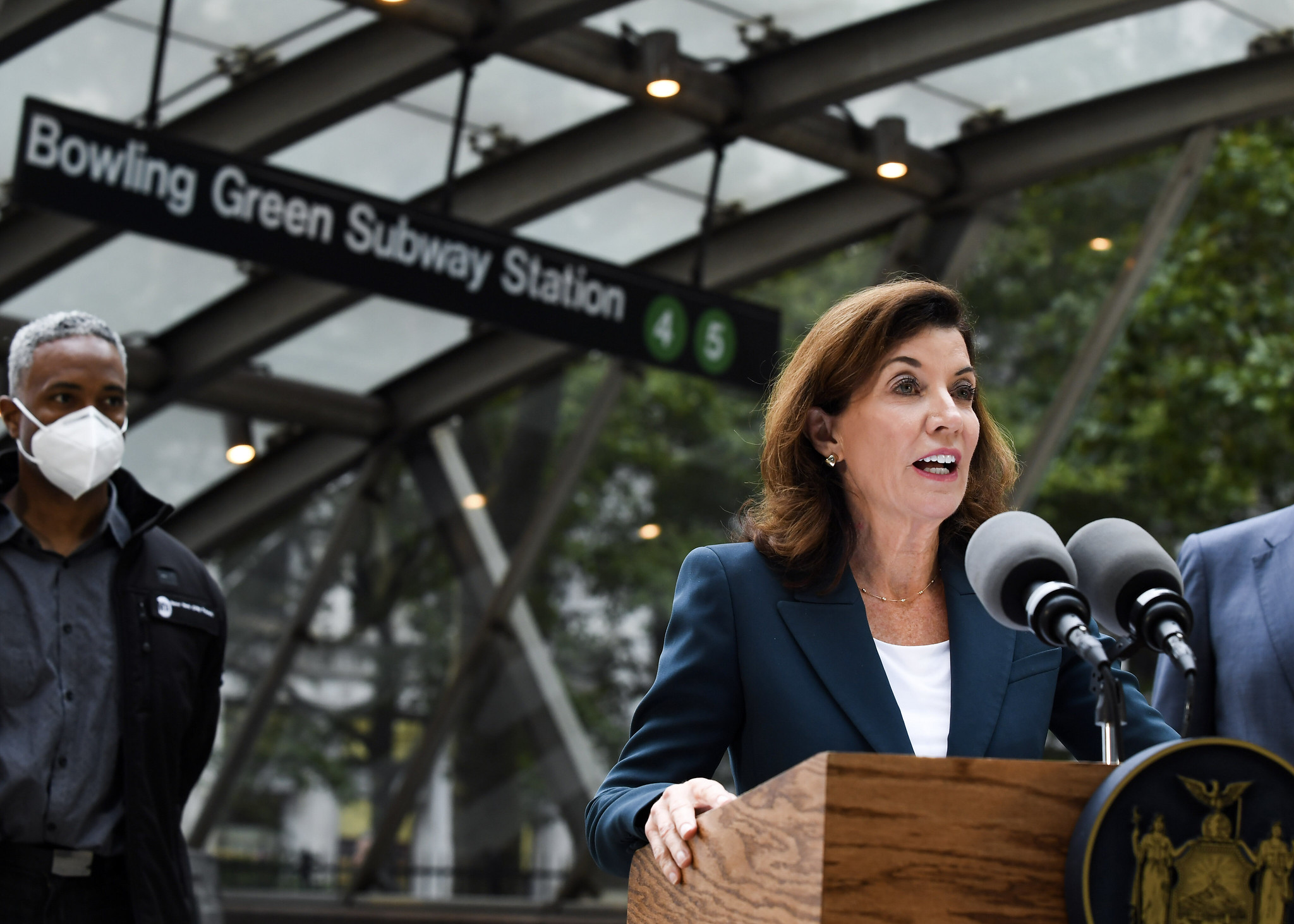 Governor Hochul, in a blue suit and white shirt, stands at a lectern outside the Bowling Greene Subway Station entrance