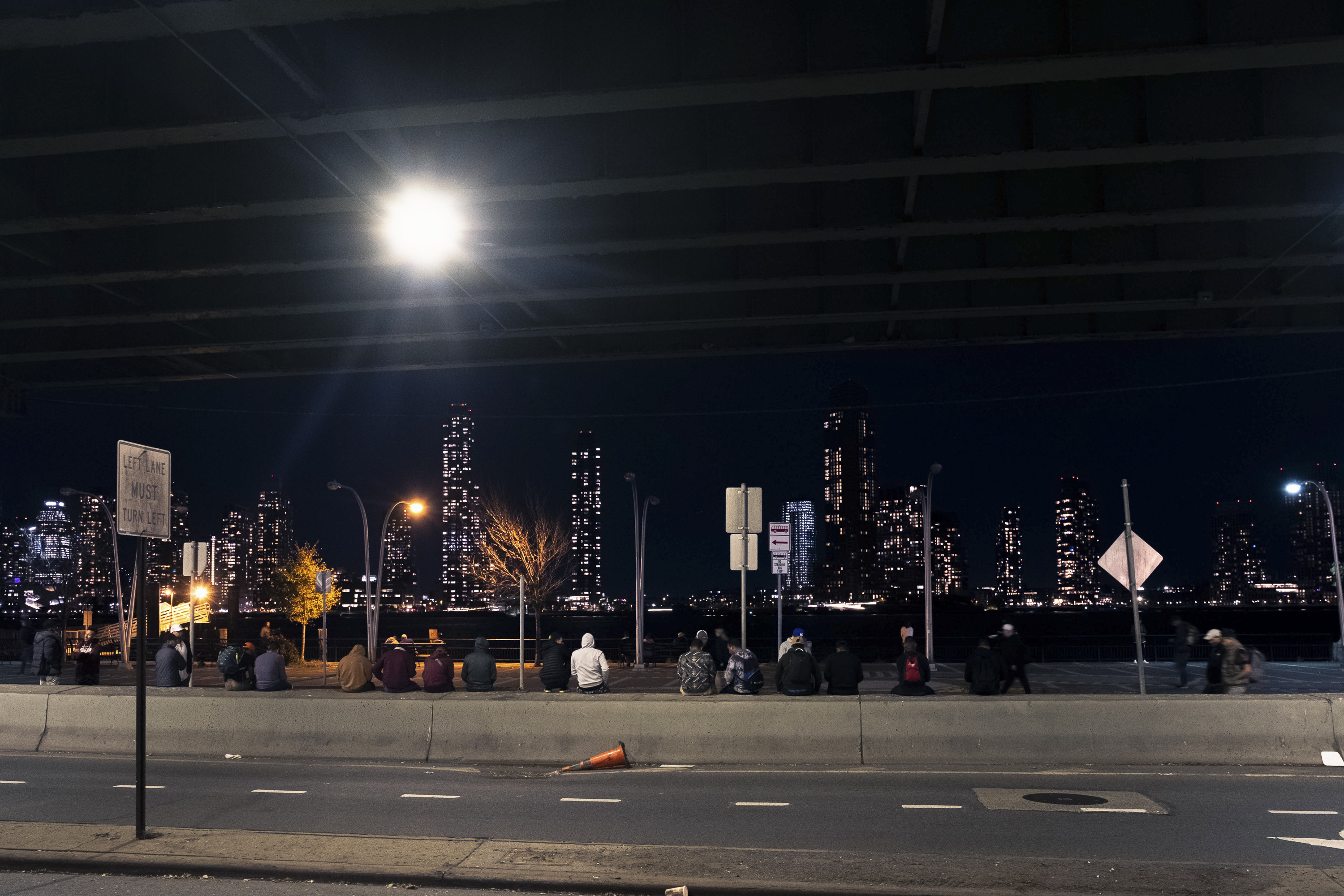 A night scene of group sitting side by side atop a divider under the FDR Drive.