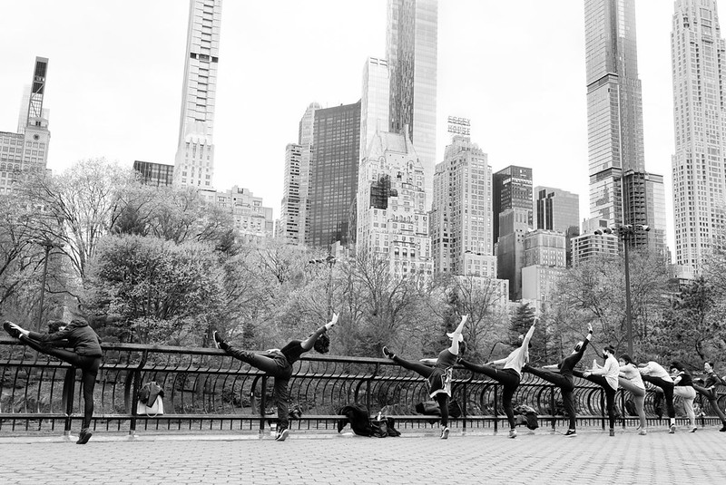 A row of dancers wearing masks and workout clothes stretch at the fence railing by Wollman Rink in Central Park.