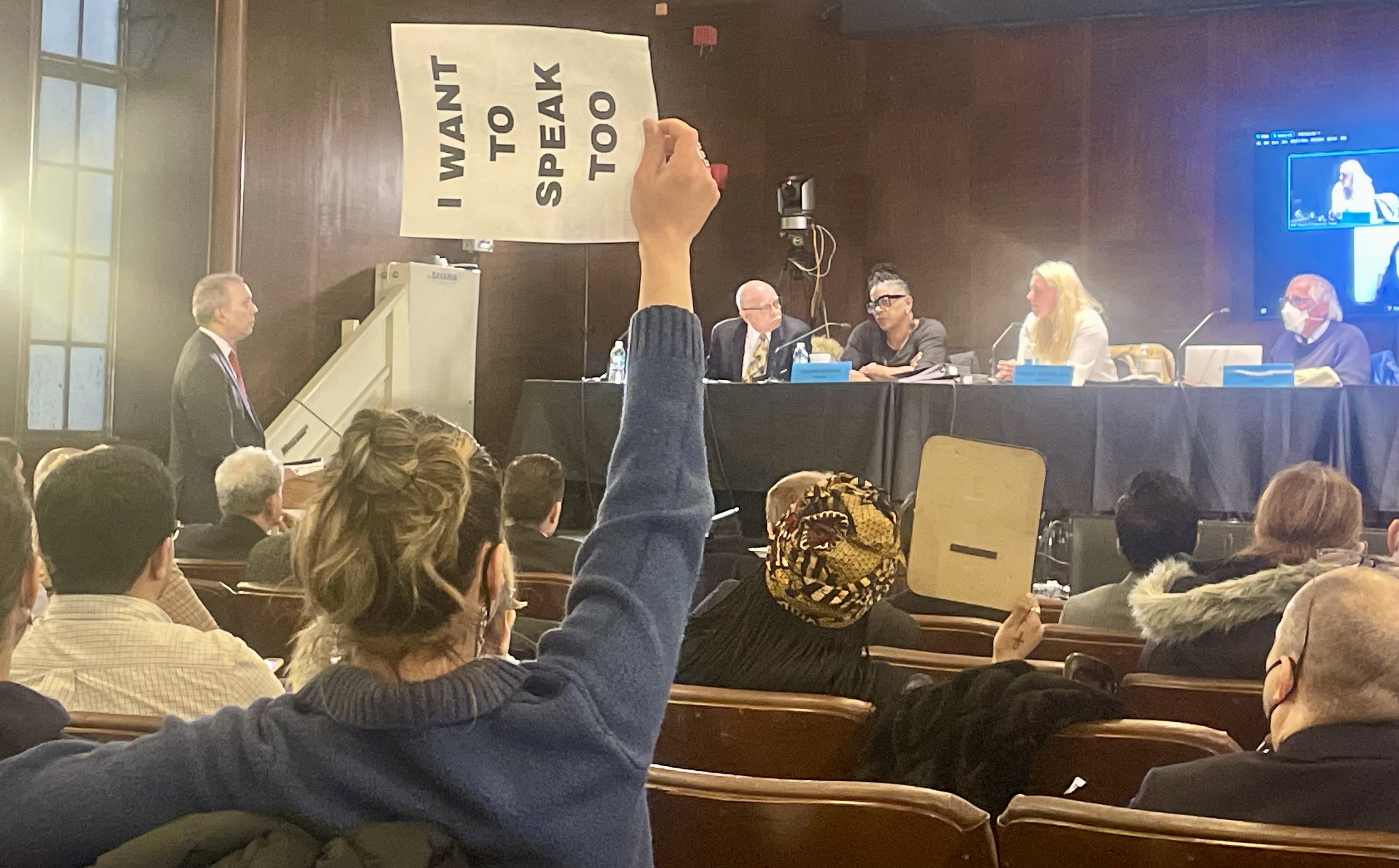 Correction Commissioner Louis Molina speaks to the Board of Correction Wednesday about his proposal to keep personal mail and packages from being sent directly to Rikers Island detainees as an activist holds up a sign protesting the board's new policy limiting the number of people who speak at its public meetings.