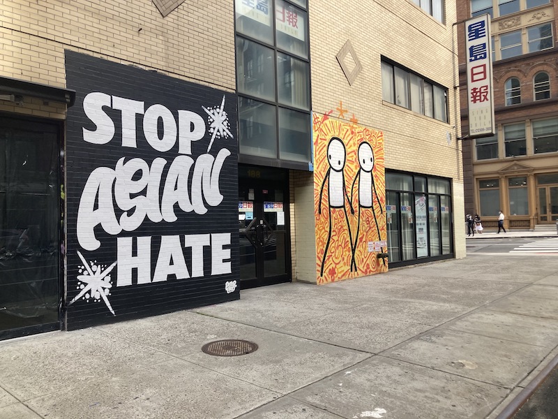 A mural that says "stop Asian hate" with two stick figures next to it adorns a wall in Chinatown as people walk by in the background.