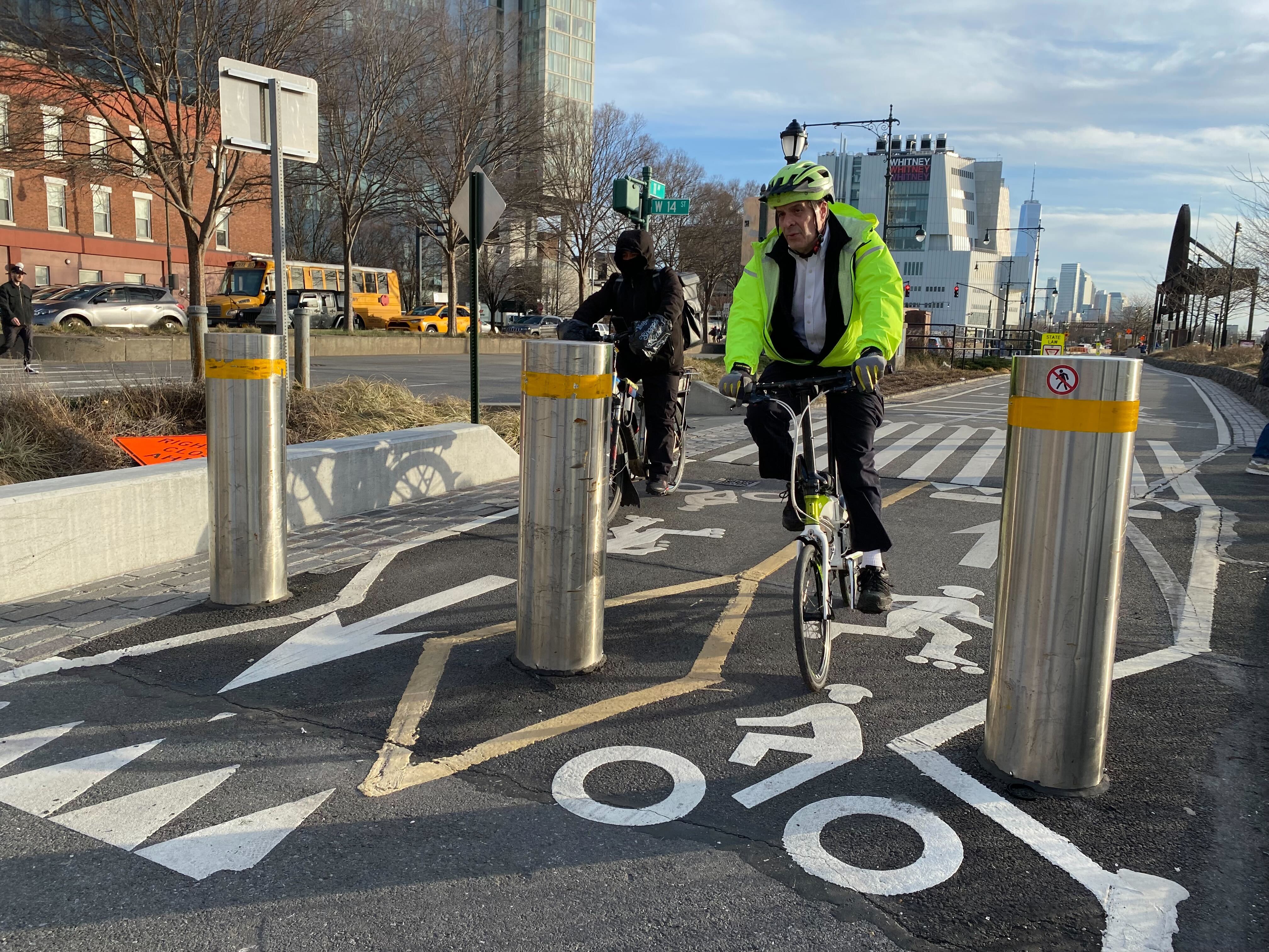 A cyclist rides between two steel bollards on a path near the Hudson River.