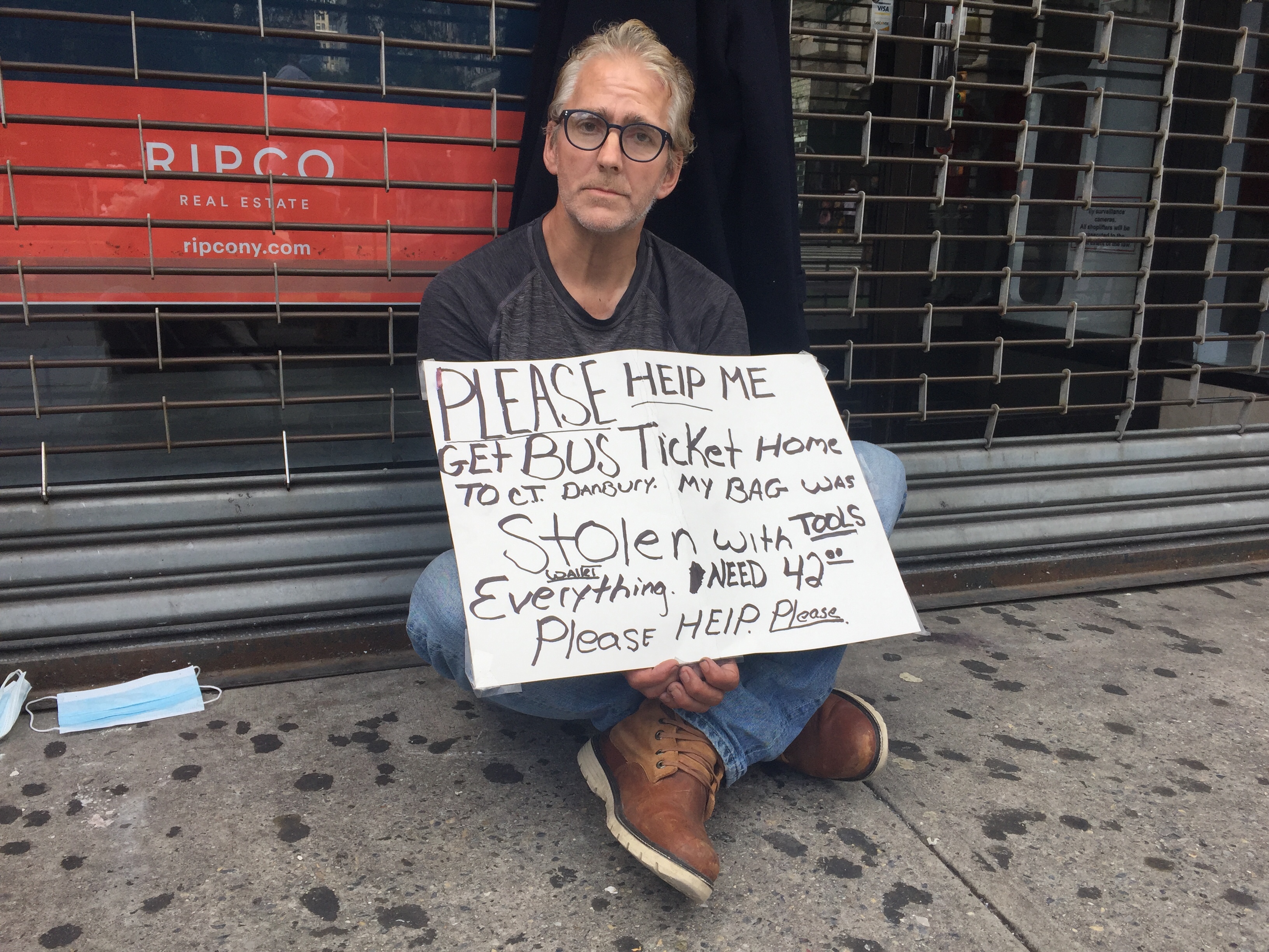 A white man with glasses, with a sign saying he's homeless, sits outside a gated store
