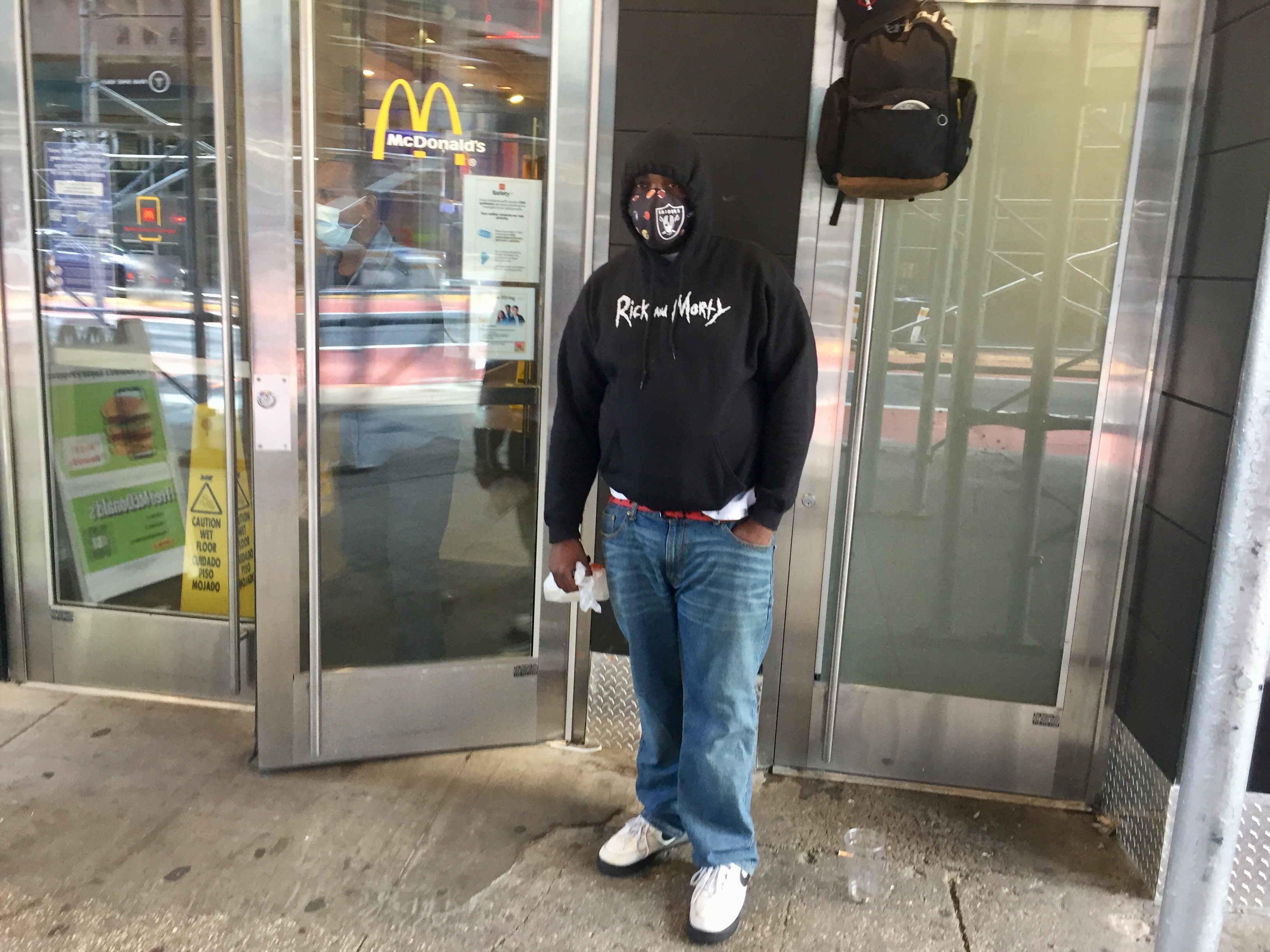 A black man wearing jeans and a hooded sweatshirt wears a mask outside a building
