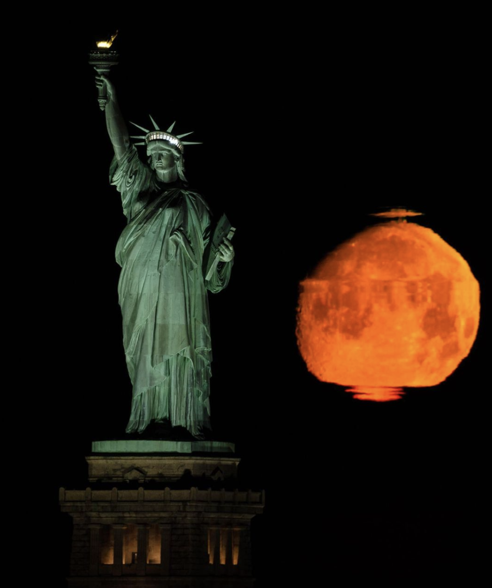 A photo of the Statue of Liberty at night next to a giant orange moon.