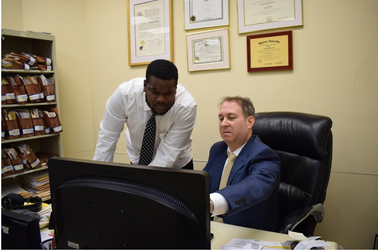 Jeremy Turnbull stands at a desk where Neill Wollerstein is sitting; both are looking at a computer.