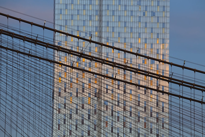 A photo of a tall glossy skyscraper rising behind the Brooklyn Bridge as seen from the South Street Seaport.