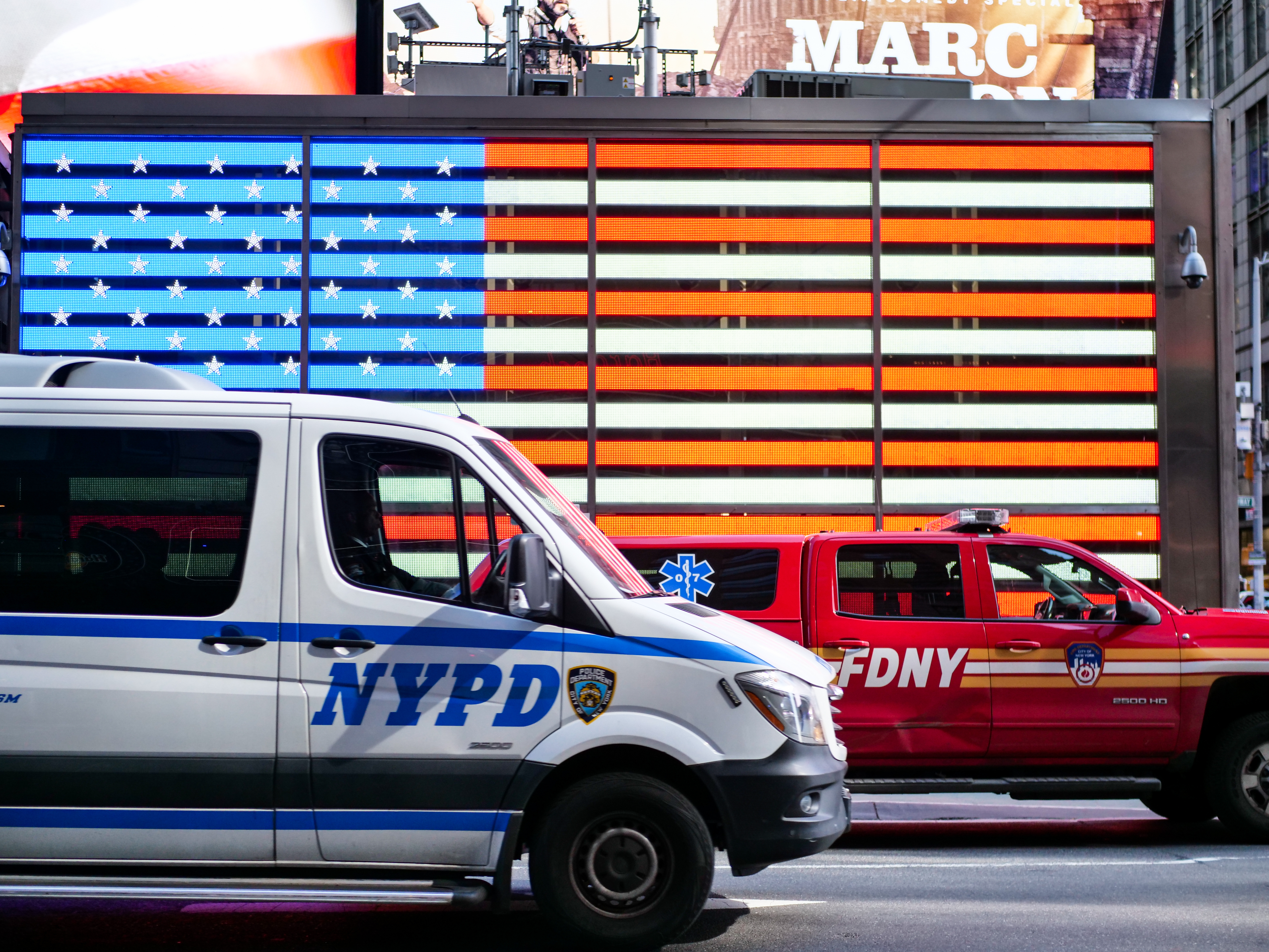 A white NYPD van drives in front of an illuminated American flag