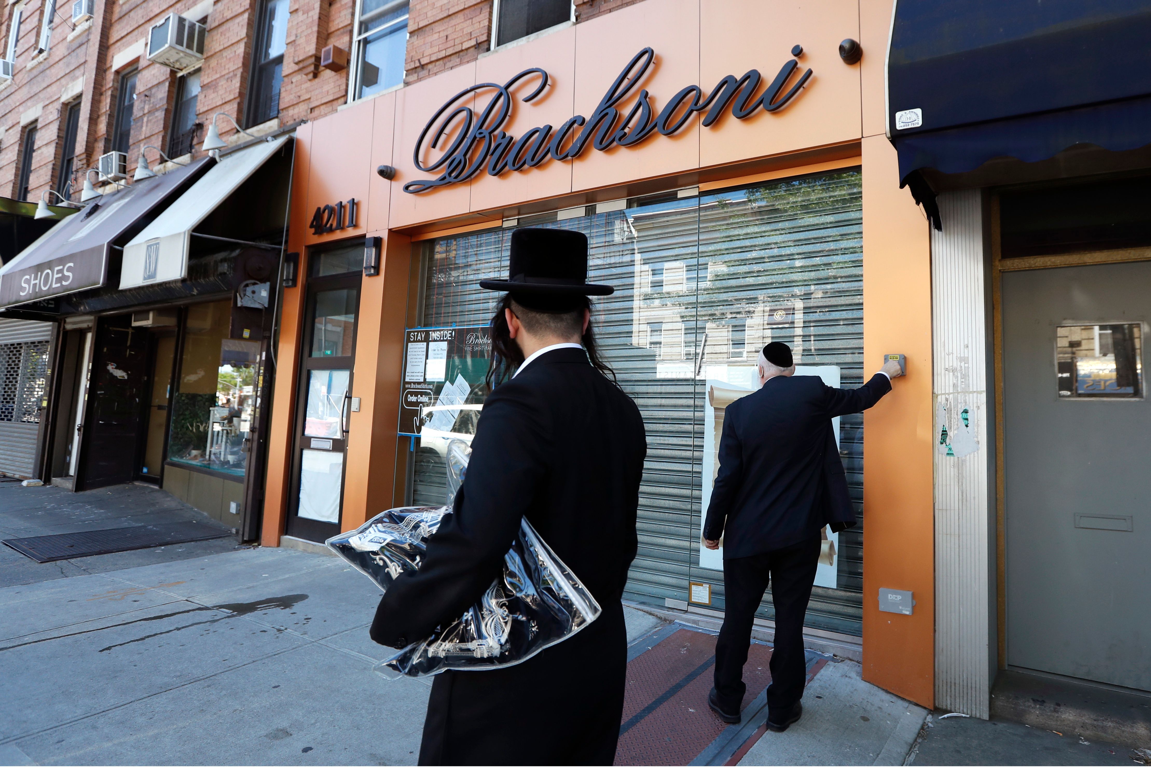Pedestrian watches as Eli Brach, right, owner of Brachsoni, which specializes in the sale of men's white shirts, raises the protective shutter on his store, in Borough Park.