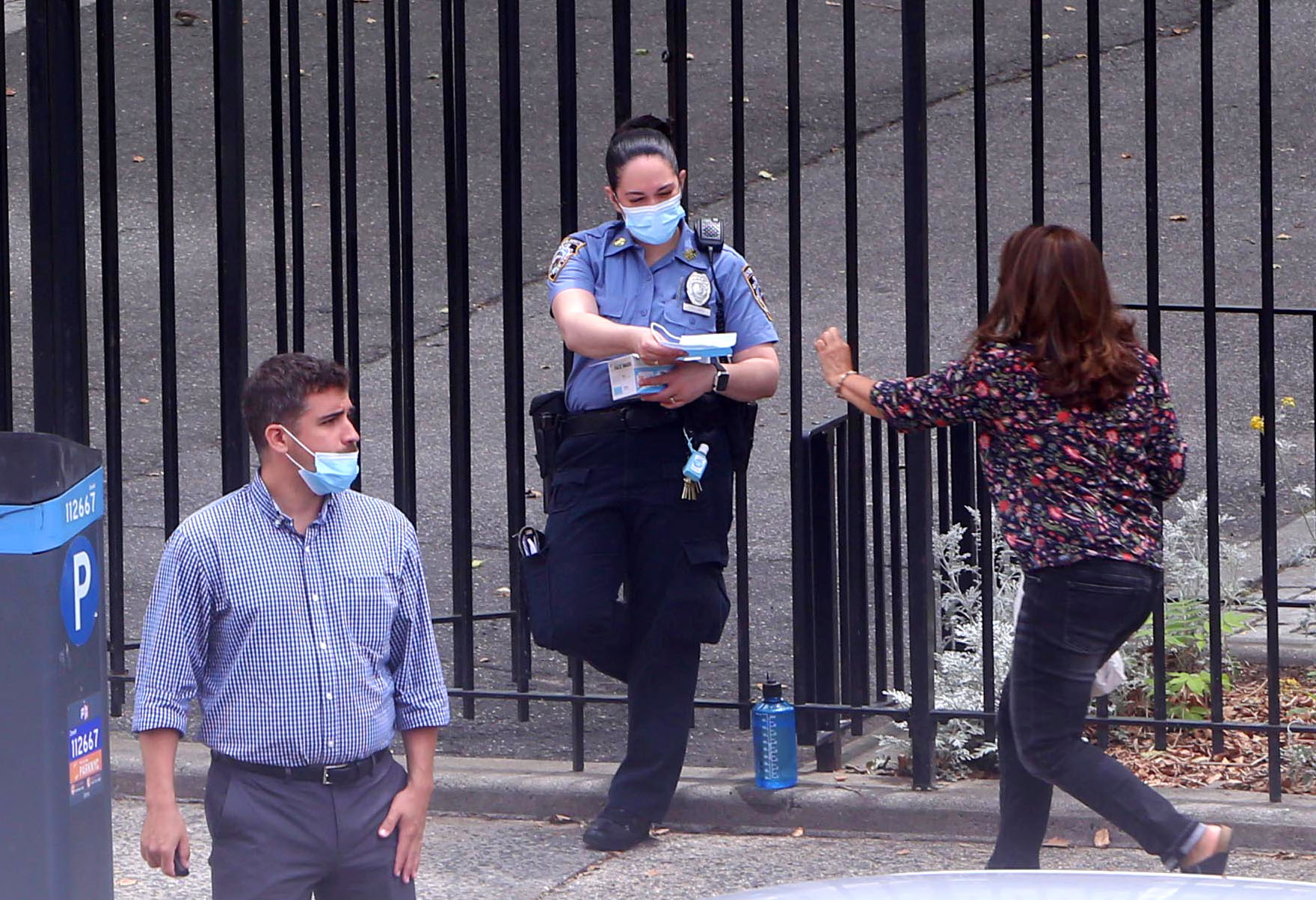 An NYPD officer in a mask hands out masks to people outside an Upper West Side Park. One man in the foreground has the mask under his nose.