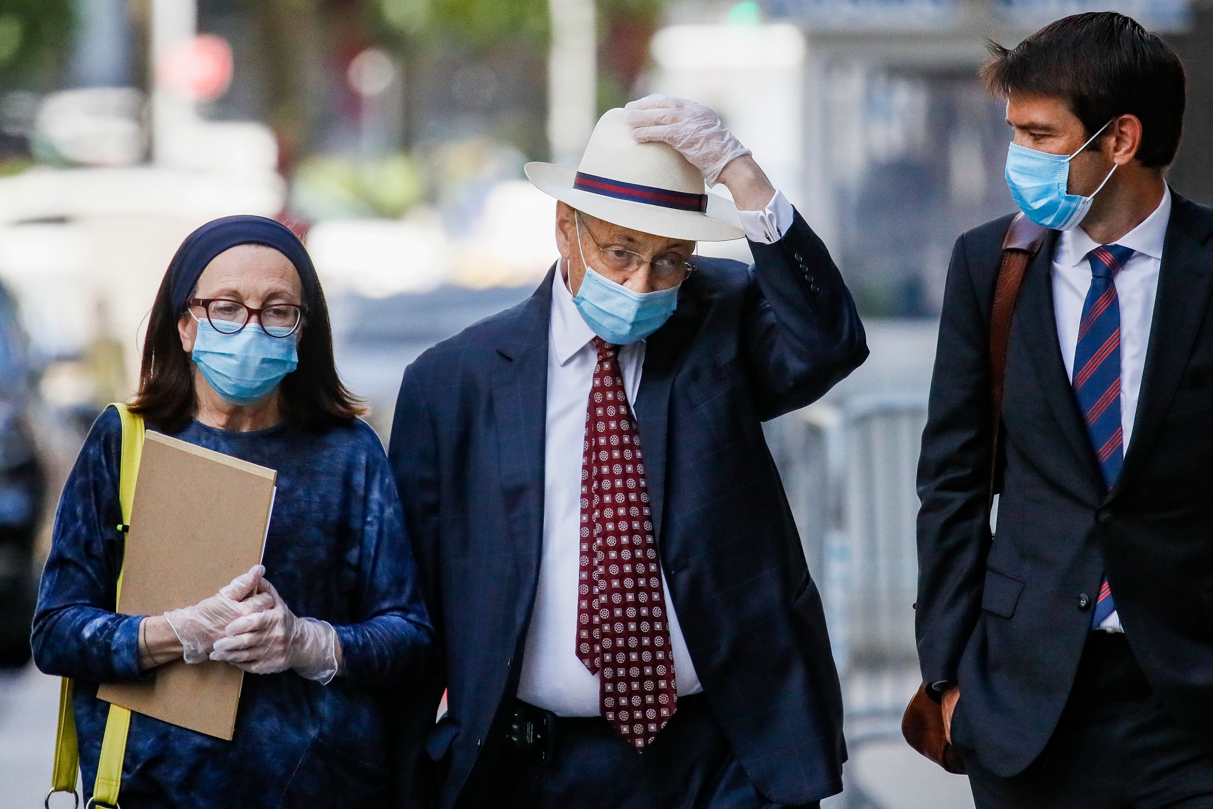 Sheldon Silver, wearing a surgical mask and gloves, wears a suit and hat while walking with two aides
