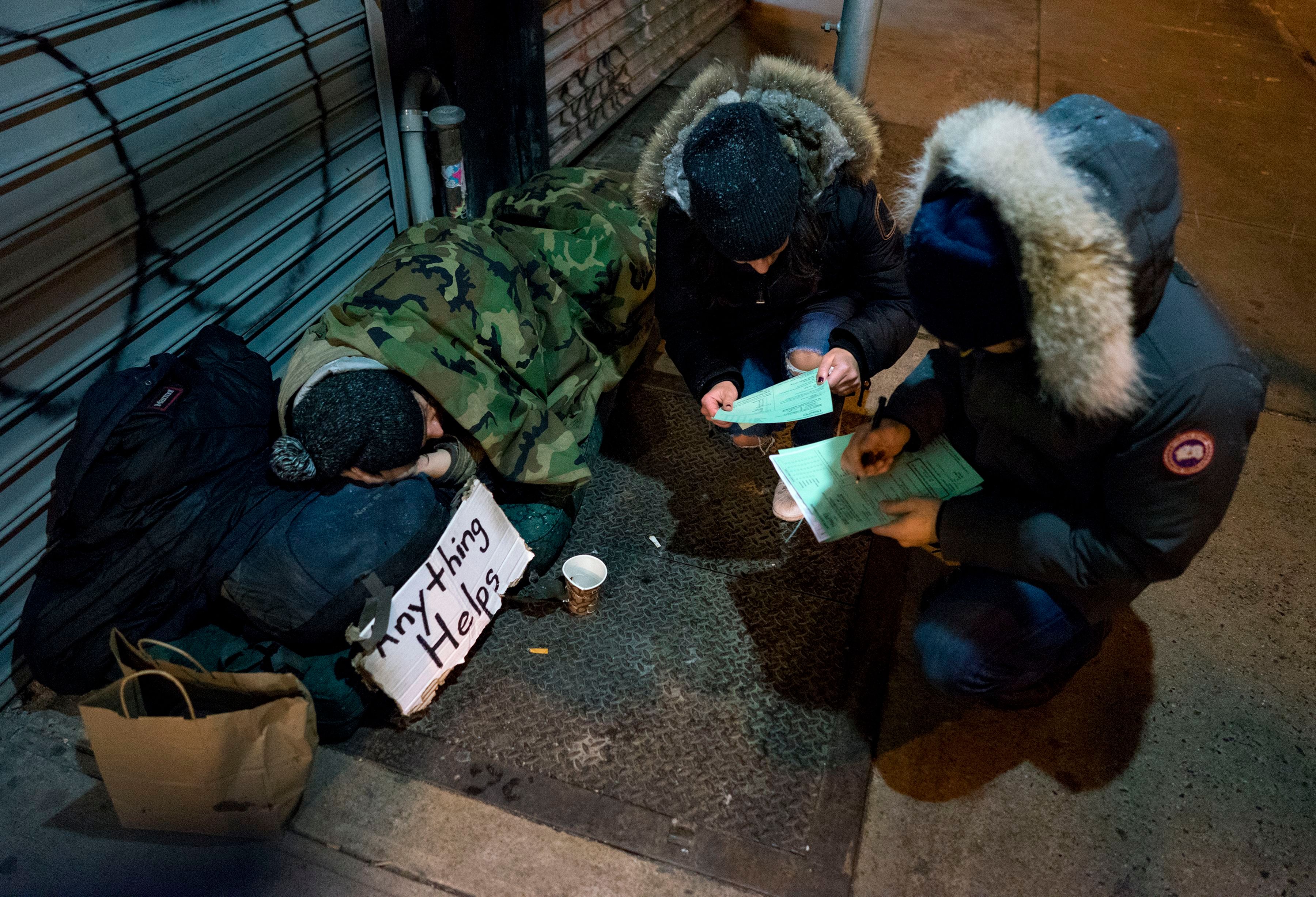 Two people look at forms while kneeling on a sidewalk near a homeless person who is resting under a blanket and next to a sign that says "Anything helps" and a donation cup